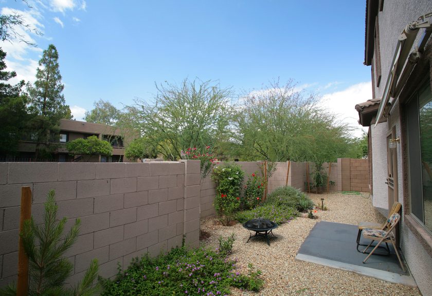 Gravel backyard closed in by block walls, with flowering shrubs, a fire pit and a small patio