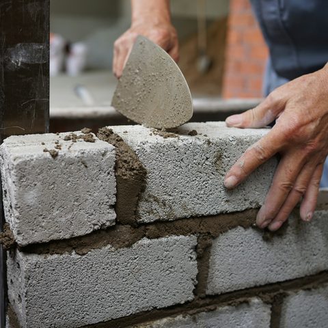 Mason pressing a gray block into place beside a fresh mortar joint, trowel in hand