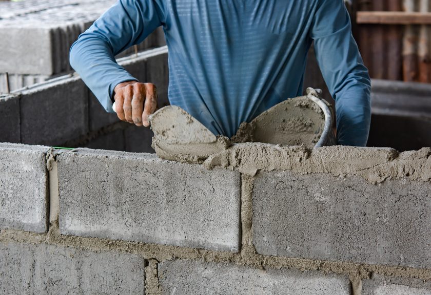 Mason spreading mortar from a trowel along the top of a gray block wall